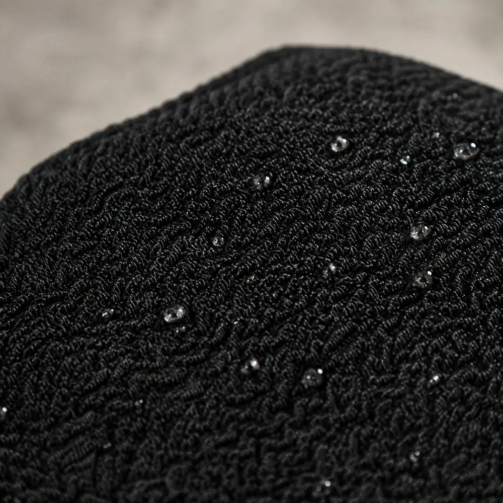 Close-up of the woven crepe texture of a black Moroccan kessa glove, with water beading on the fabric
