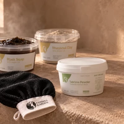 The Fatima's Garden hammam set: black soap with argan oil, rhassoul clay with argan oil, traditional tabrima powder and a black kessa glove, on a stone ledge